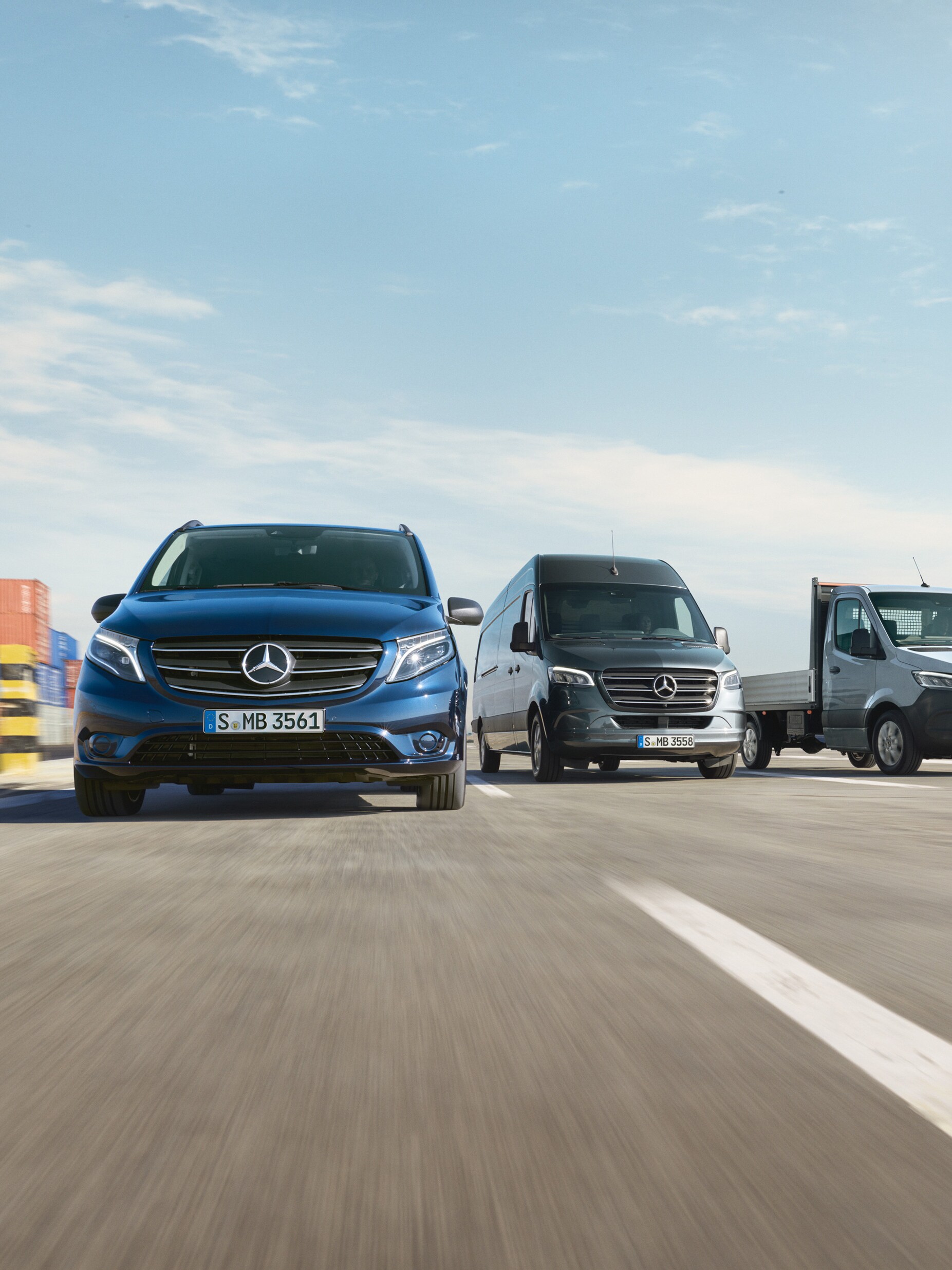 A range of Mercedes-Benz vans parked side-by-side on a road next to a shipping dock.