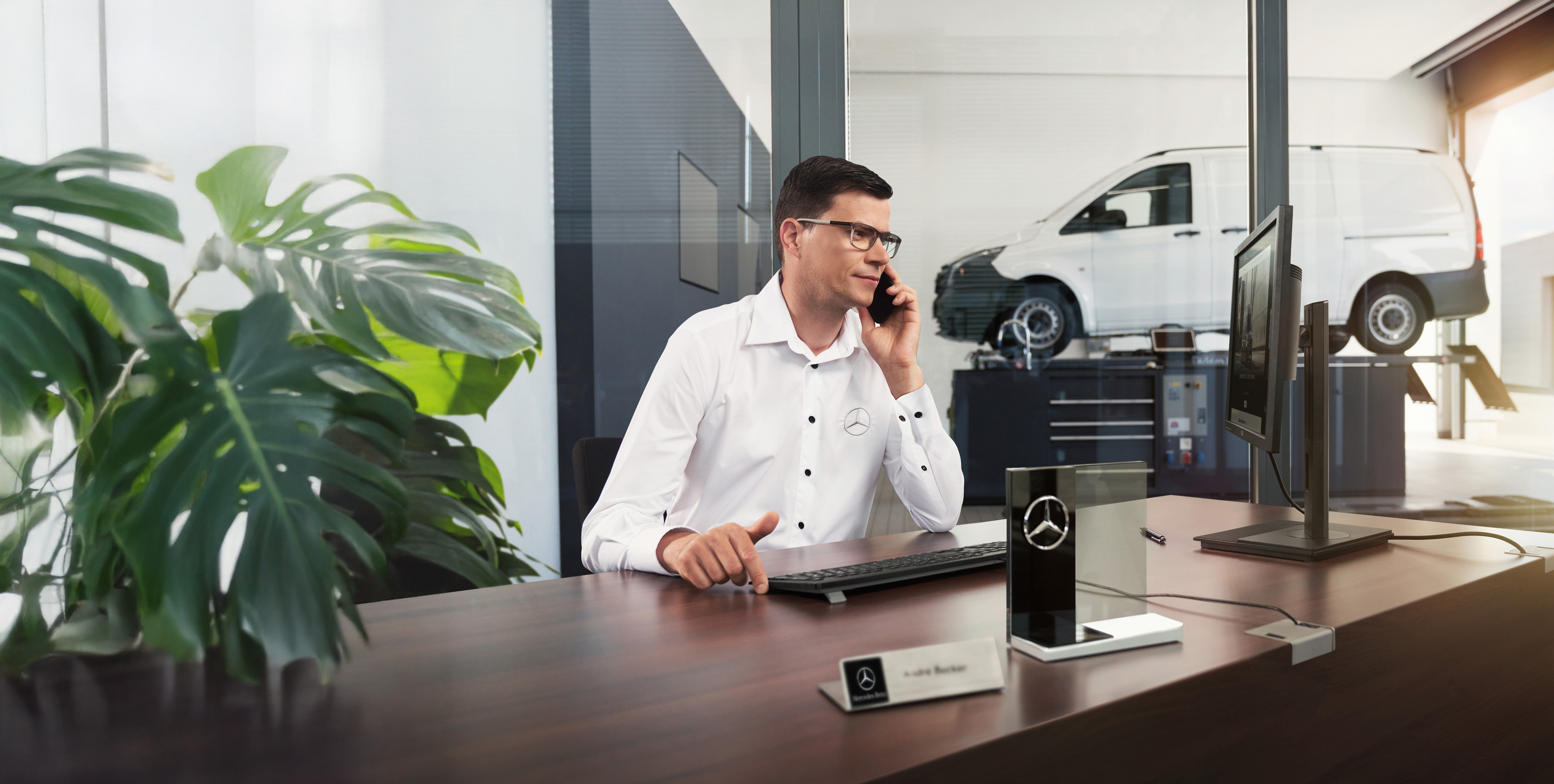 Mercedes-Benz | Vans Care Mercedes-Benze employee talks on the phone at a desk with a Mercedes-Benz Vito van in the workshop.
