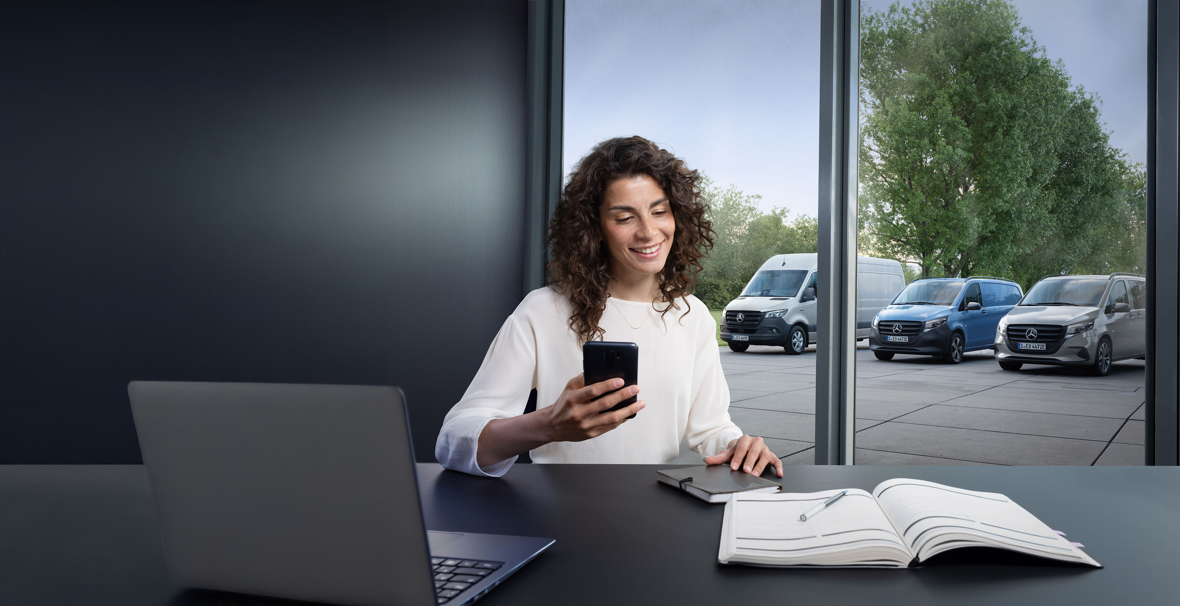 Mercedes-Benz | Convenience | Authorised Repairer Woman looking for a Mercedes-Benz Authorised Repairer on her smartphone with a range of Mercedes-Benz vans in the background.