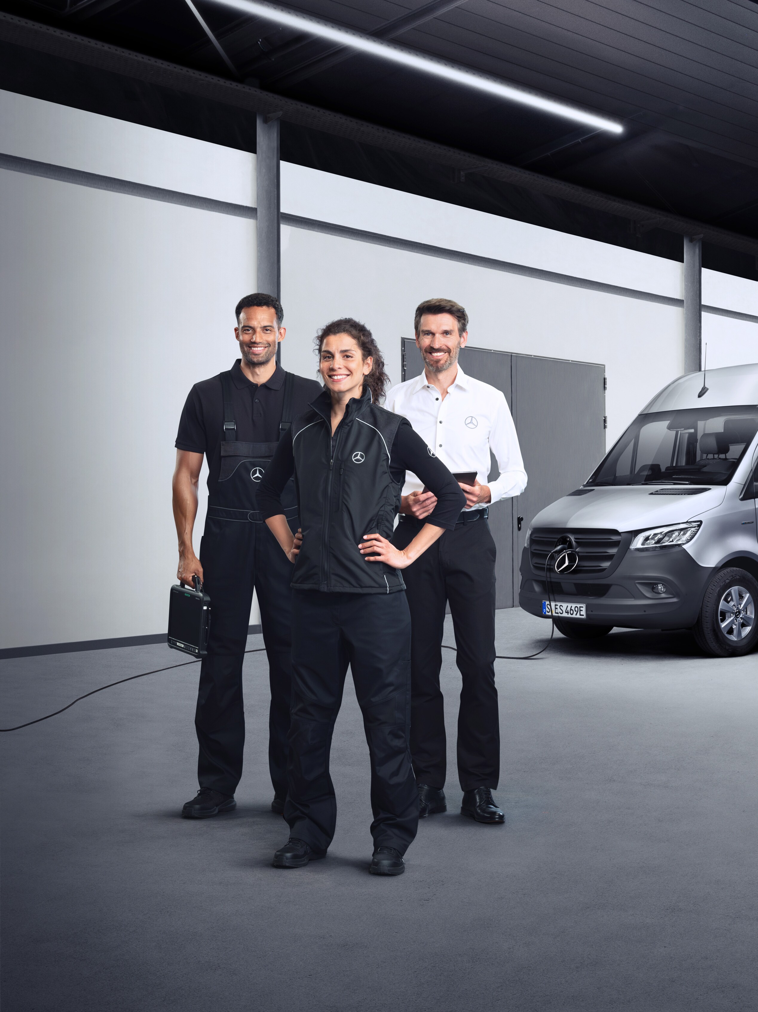 Three employees in front of an electric Mercedes-Benz Sprinter van with charging cable attached.