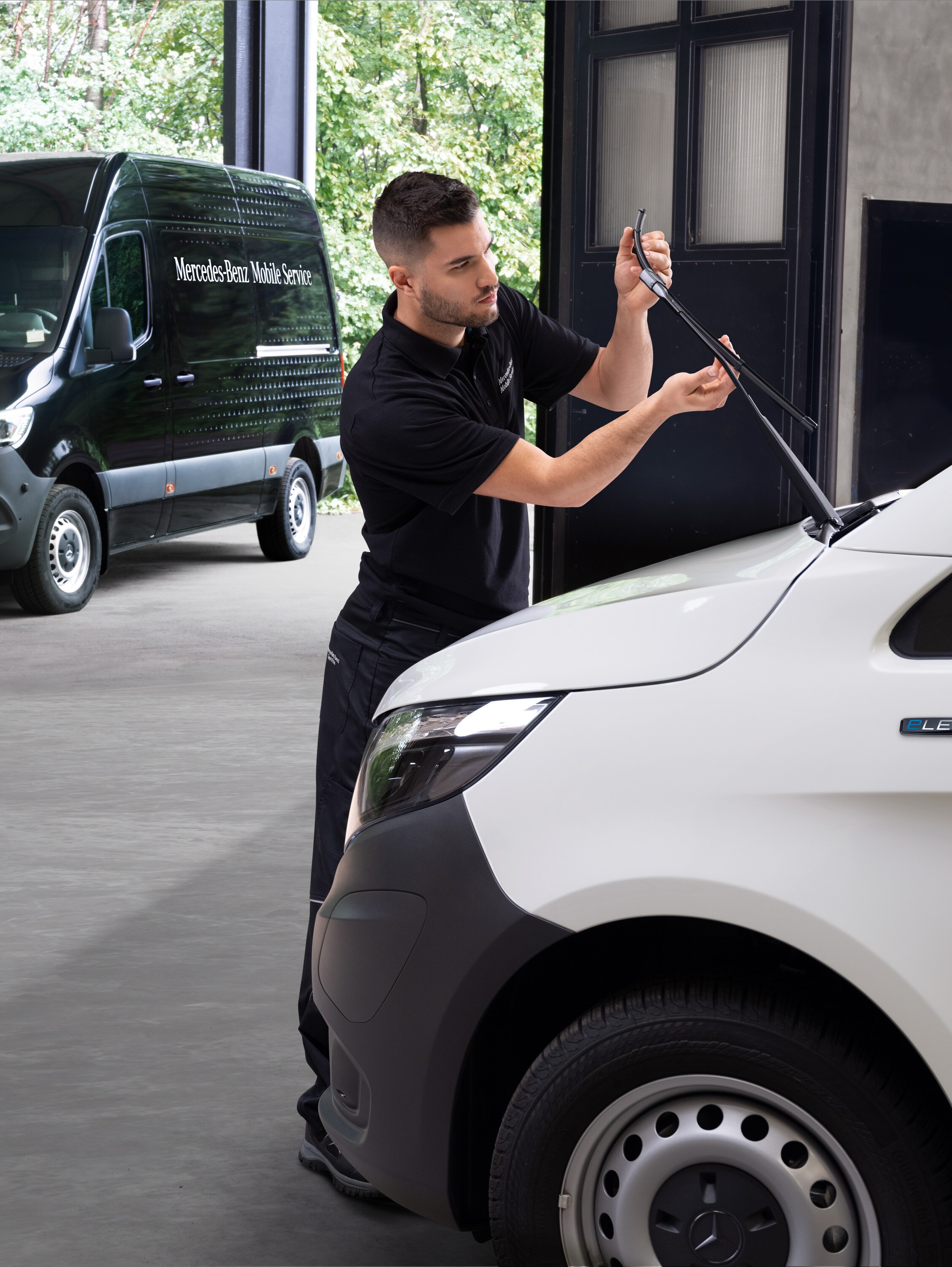 A technician changes wiper blades on a white Mercedes-Benz Vito van.