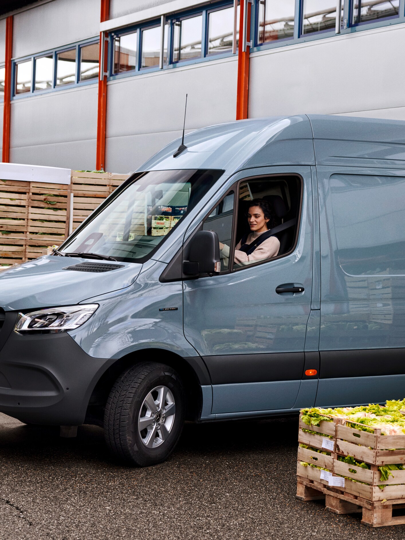 A woman sitting in the driver's seat of a grey Mercedes-Benz eSprinter van next to crates of lettuce.